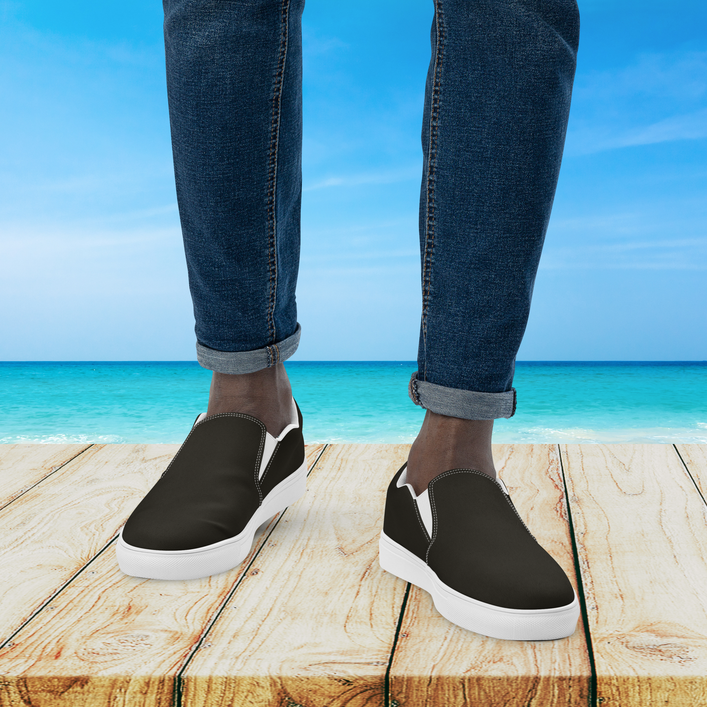Person wearing black slip-on shoes with white soles on a wooden platform by the ocean.