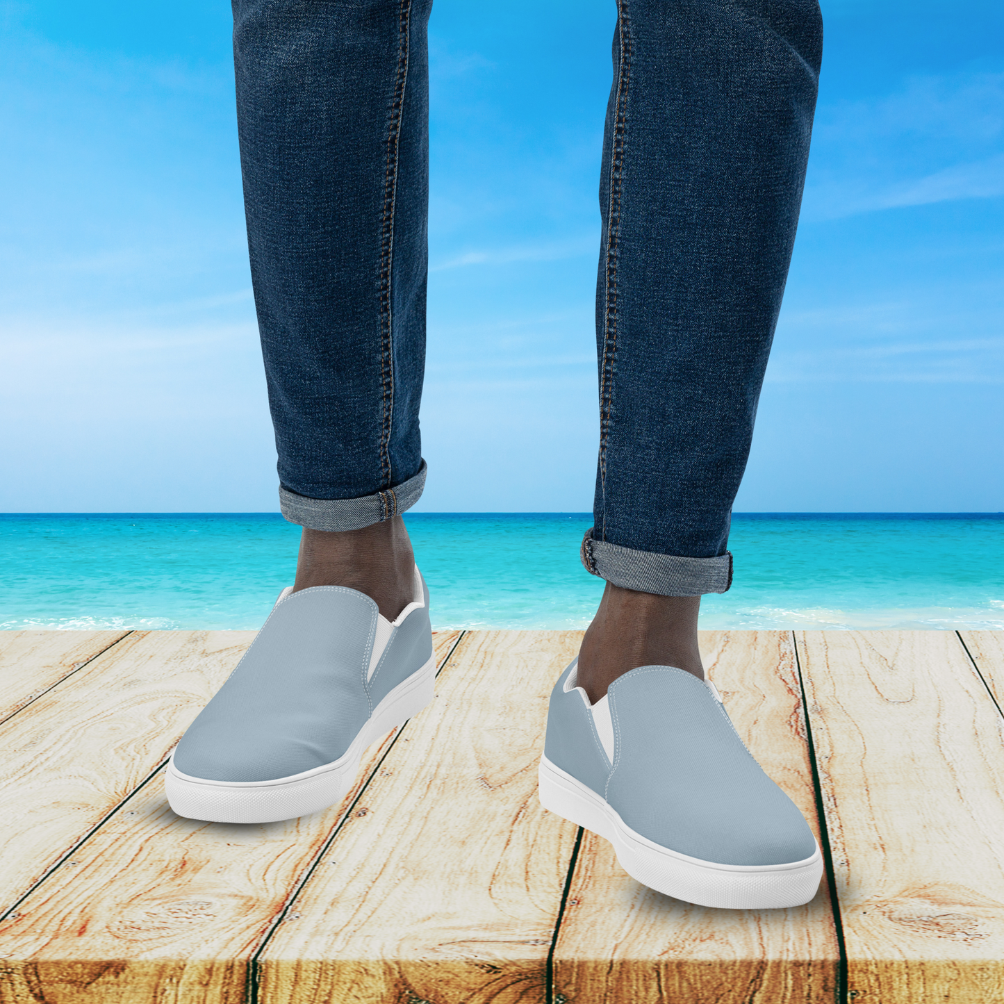 Person wearing light gray slip-on shoes with white soles on a wooden platform by the ocean.