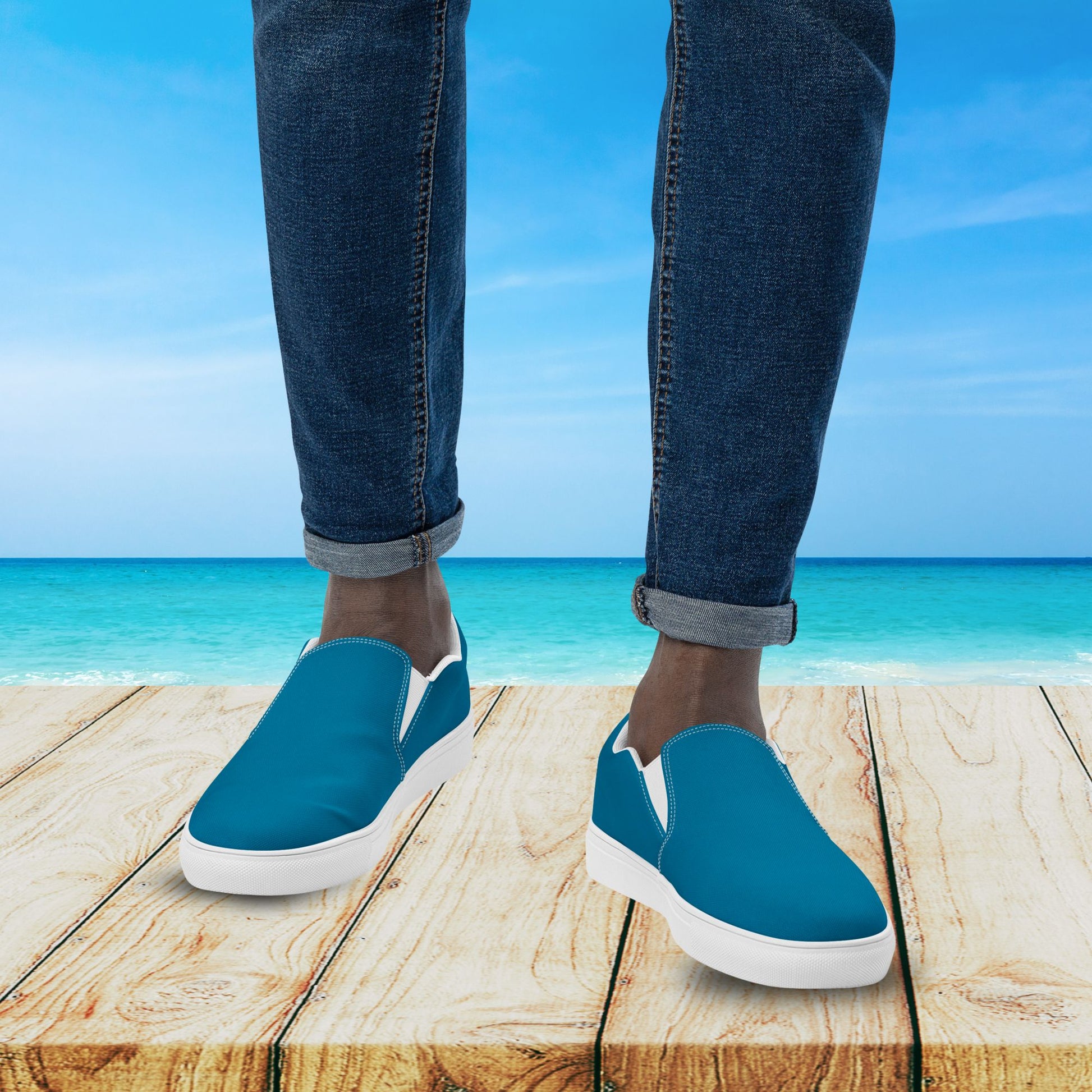 Blue slip-on shoes with white soles worn by a person standing on a wooden platform with a beach background.