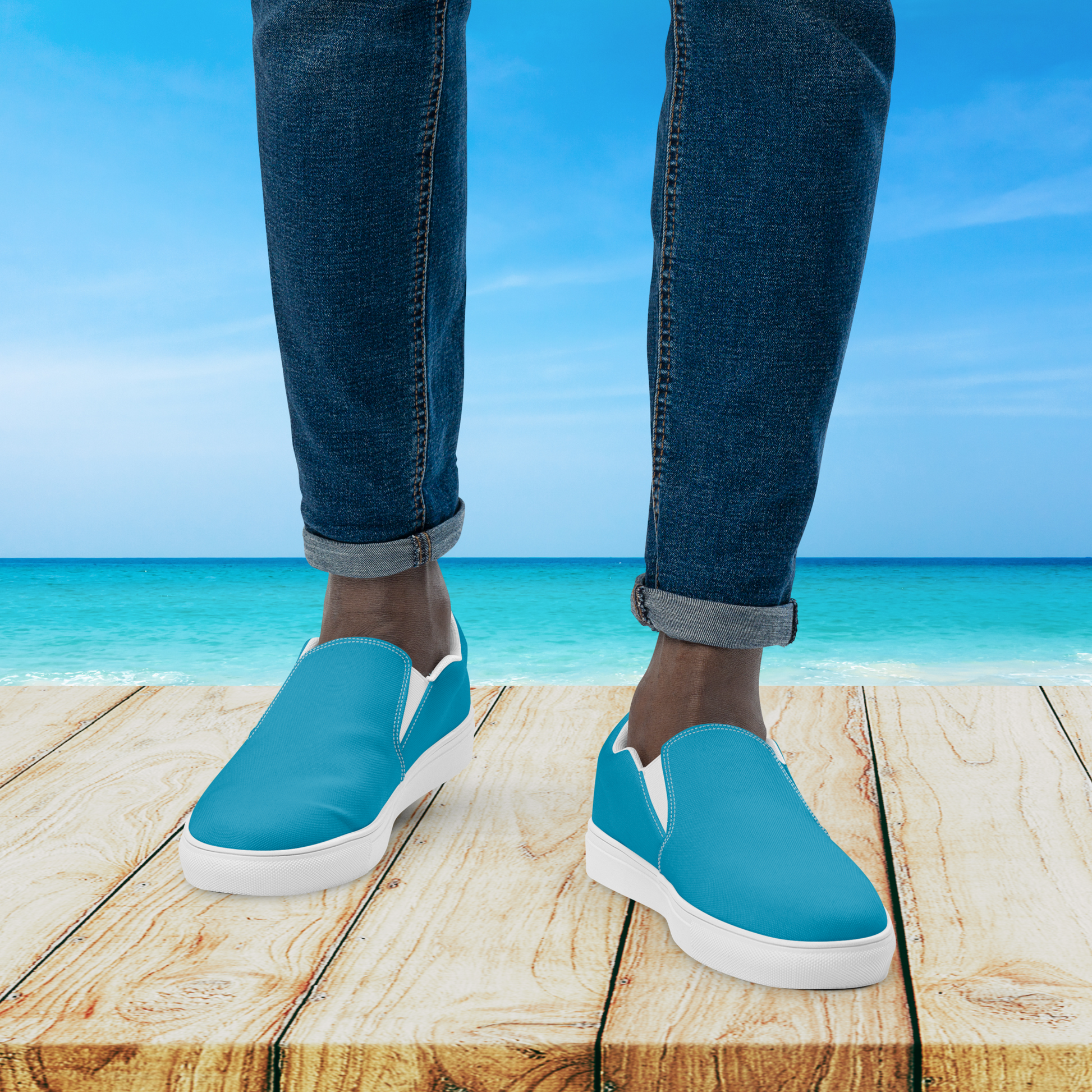 Blue slip-on shoes with white soles worn by a person standing on a wooden platform with a beach background.