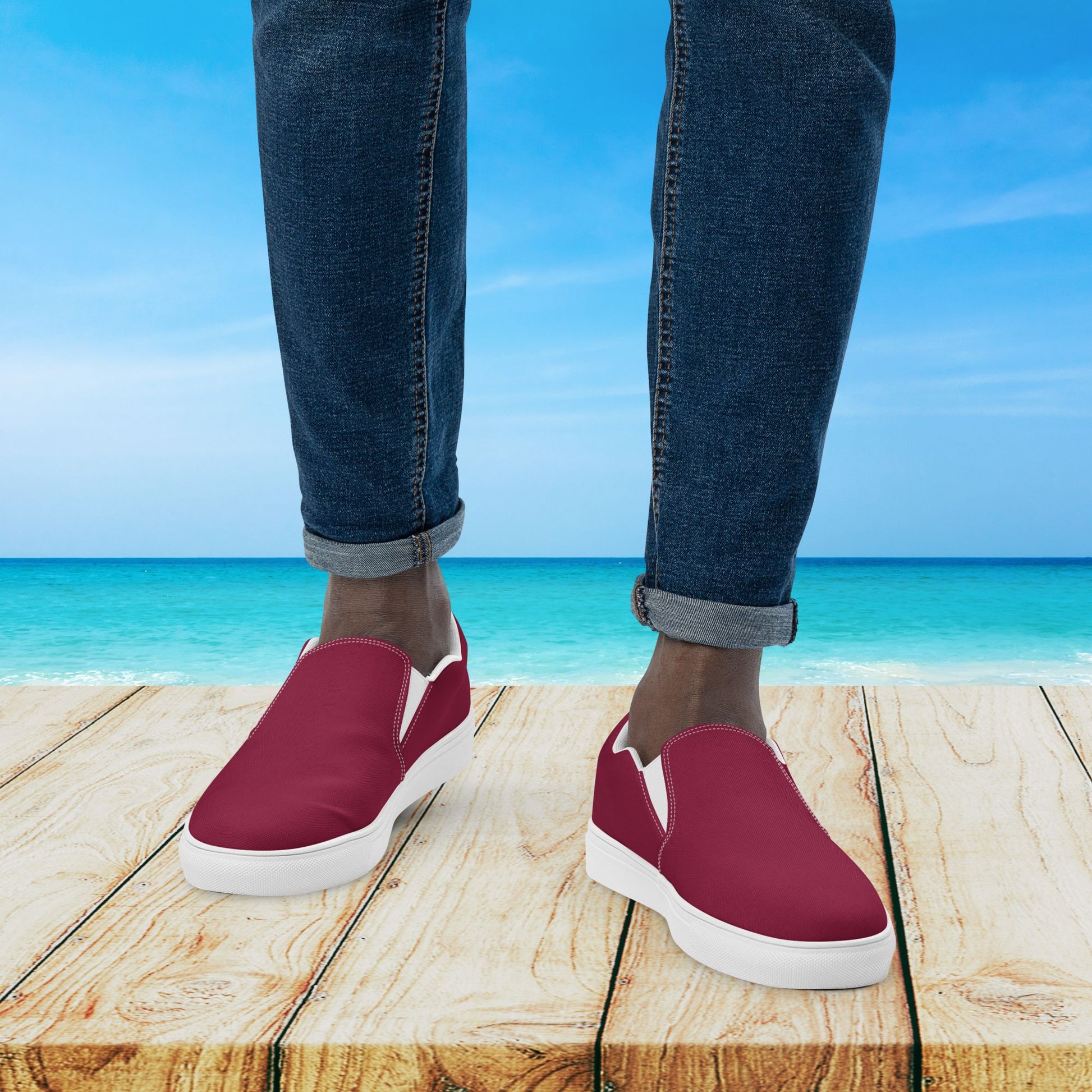 Red slip-on shoes with white soles worn by a person standing on a wooden platform with a beach background.