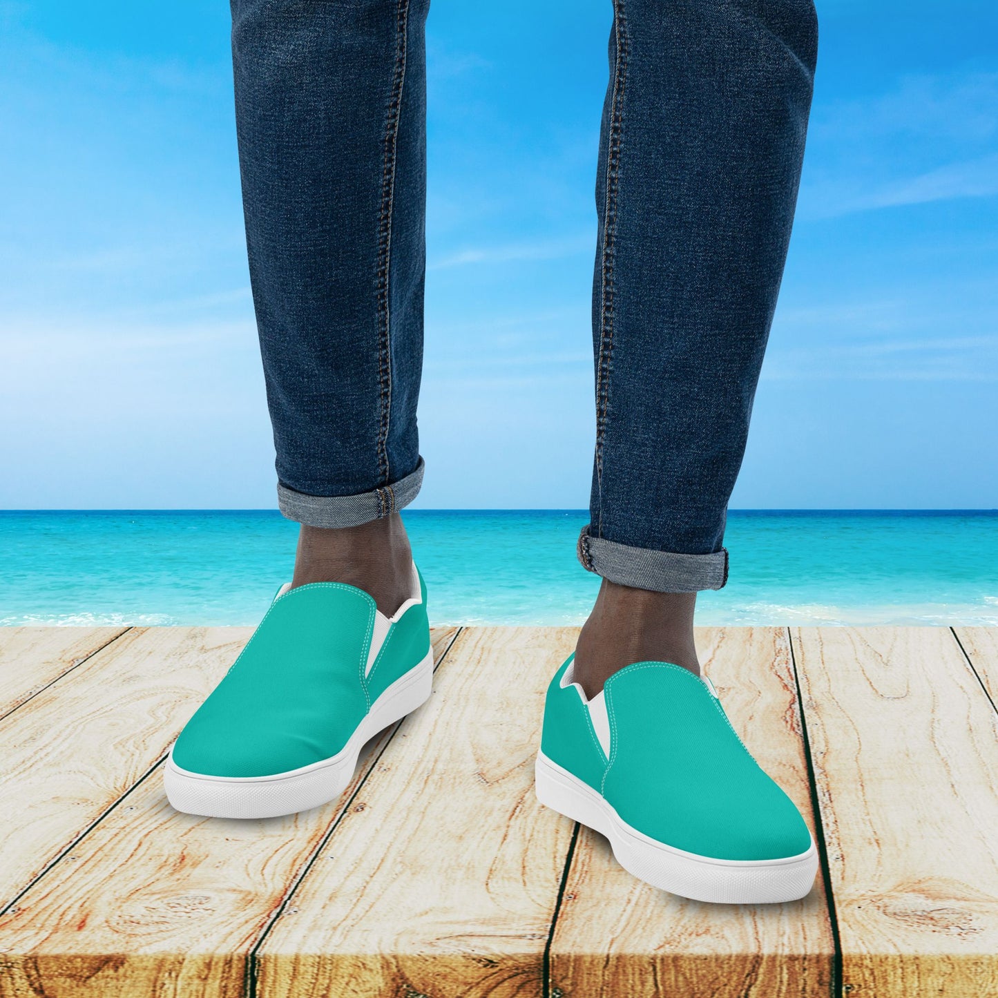 Teal slip-on shoes worn with blue jeans on a wooden platform with a beach background