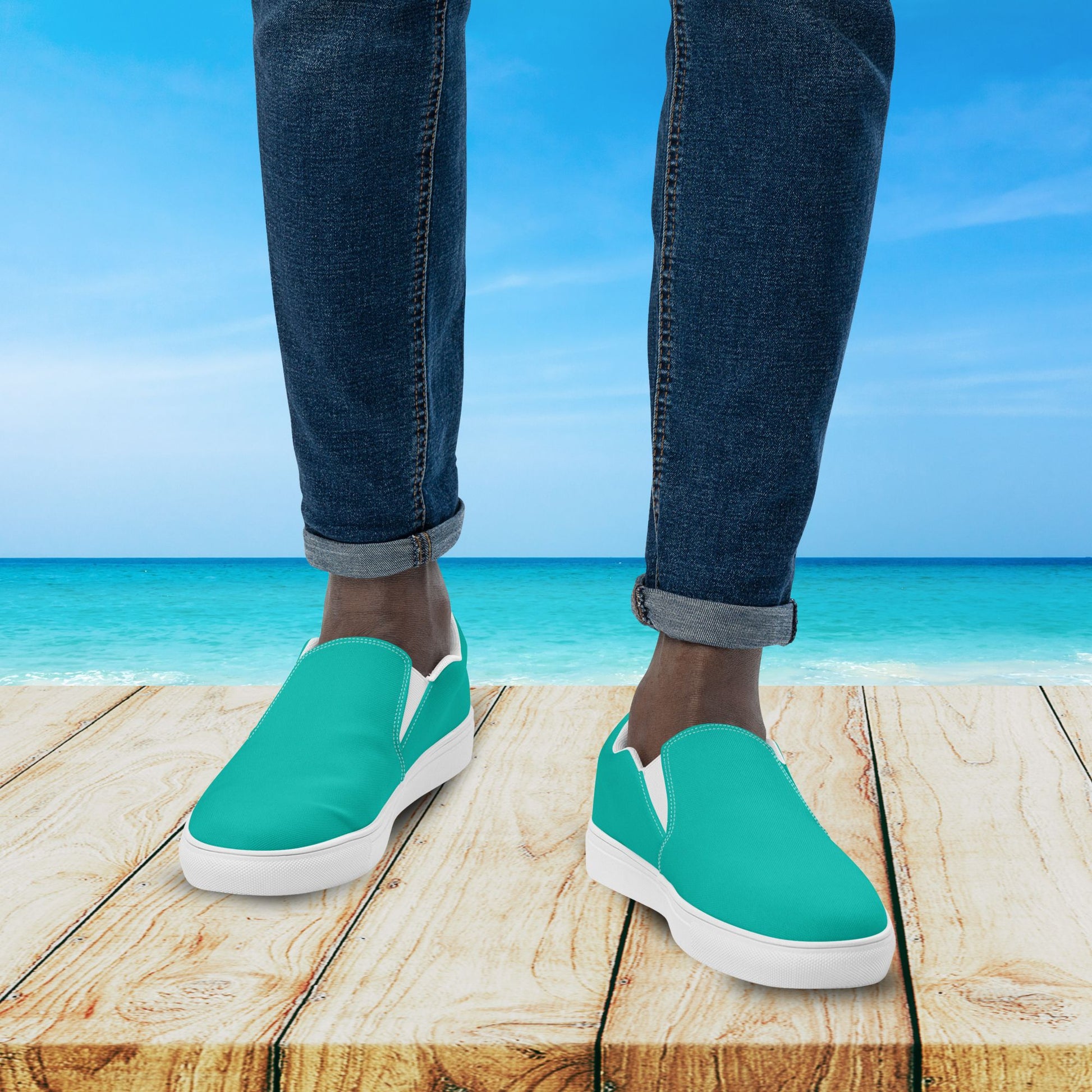 Teal slip-on shoes worn with blue jeans on a wooden platform with a beach background