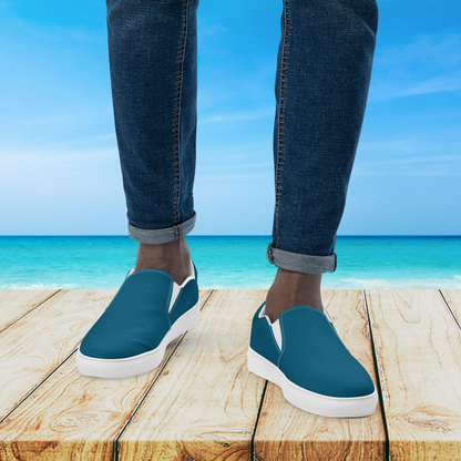 Blue slip-on shoes with white soles worn by a person standing on a wooden platform with a beach background.