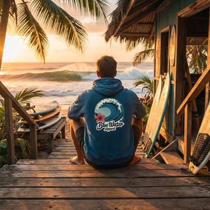 man wearing antique sapphire hibiscus wave adult hoodie sitting on steps of surf shack overlooking beach by blue water apparel