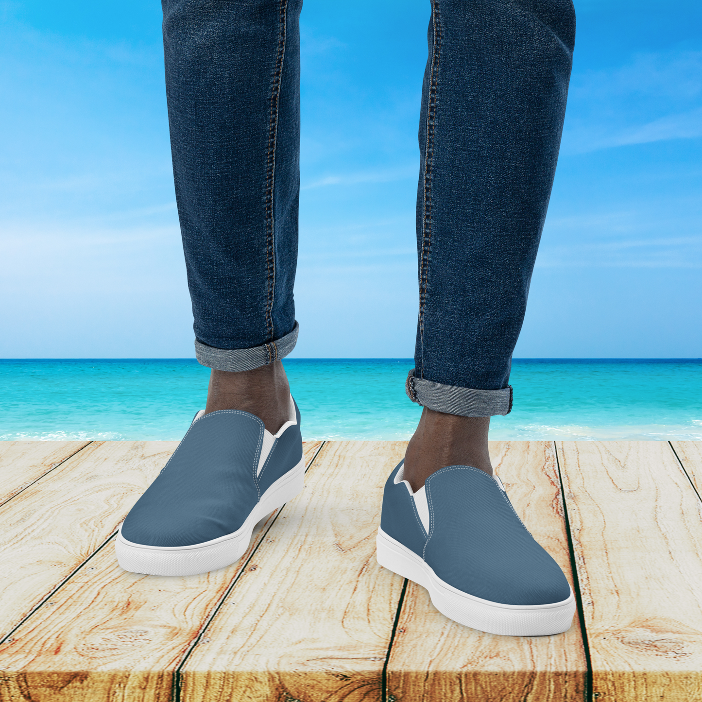 Person wearing blue slip-on shoes with white soles on a wooden platform by the ocean.