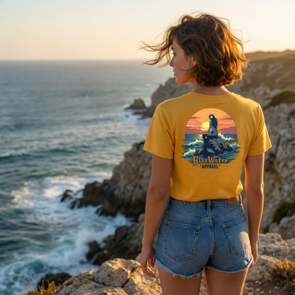 woman wearing a gold mermaid sunset tshirt standing on ocean cliff by blue water apparel