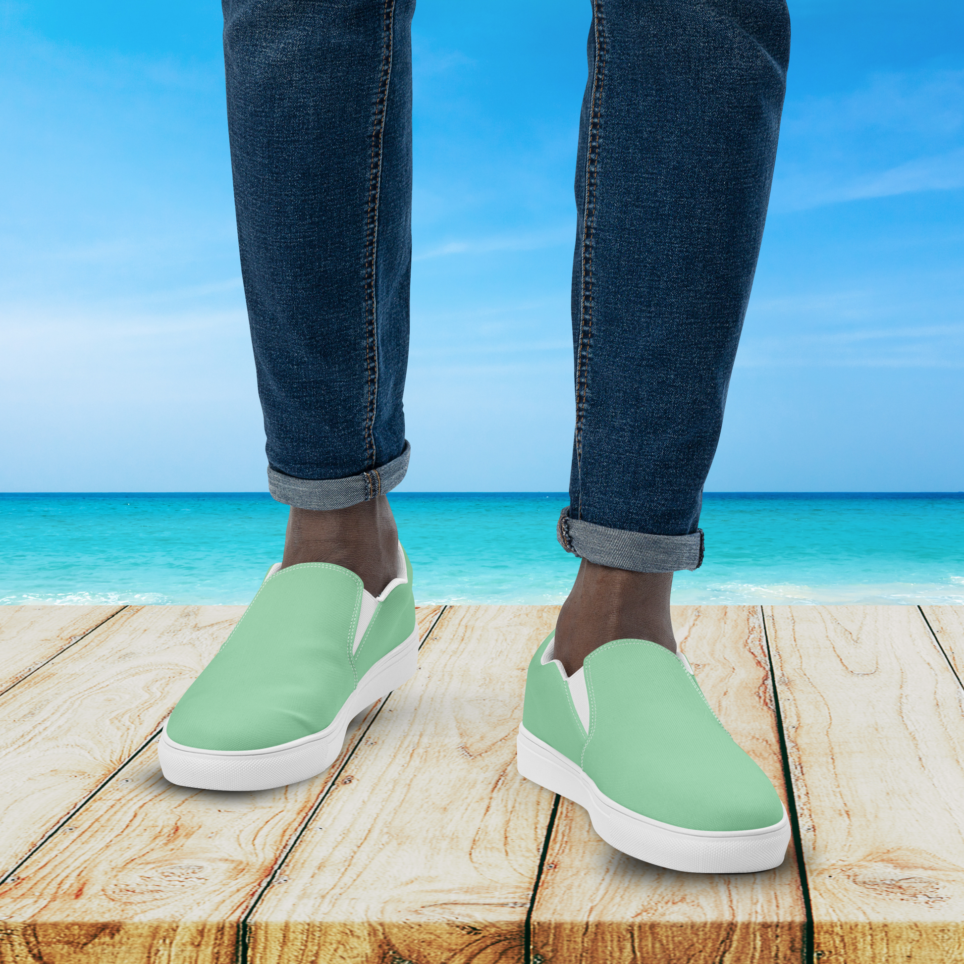 Person wearing green slip-on shoes with blue jeans standing on a wooden platform by the ocean.