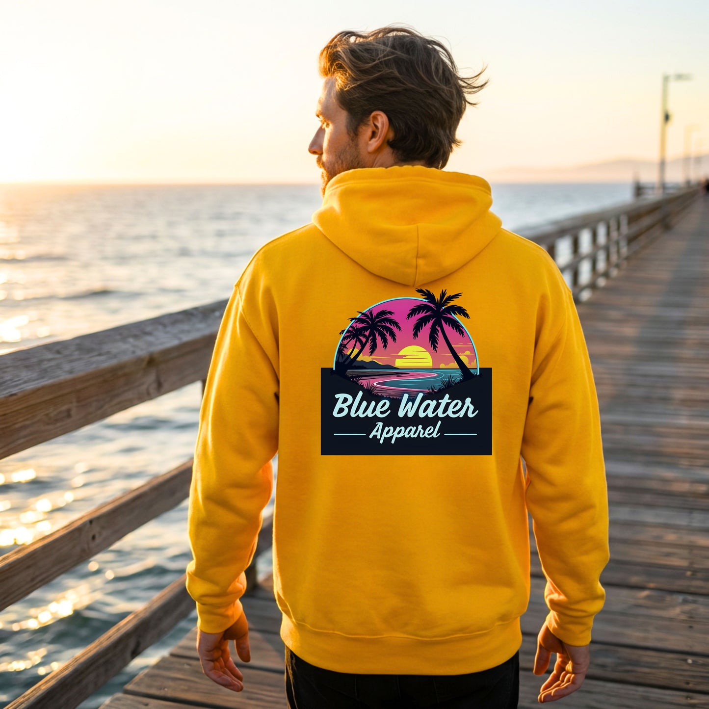 male model wearing gold neon sunset adult hoodie on fishing pier by blue water apparel