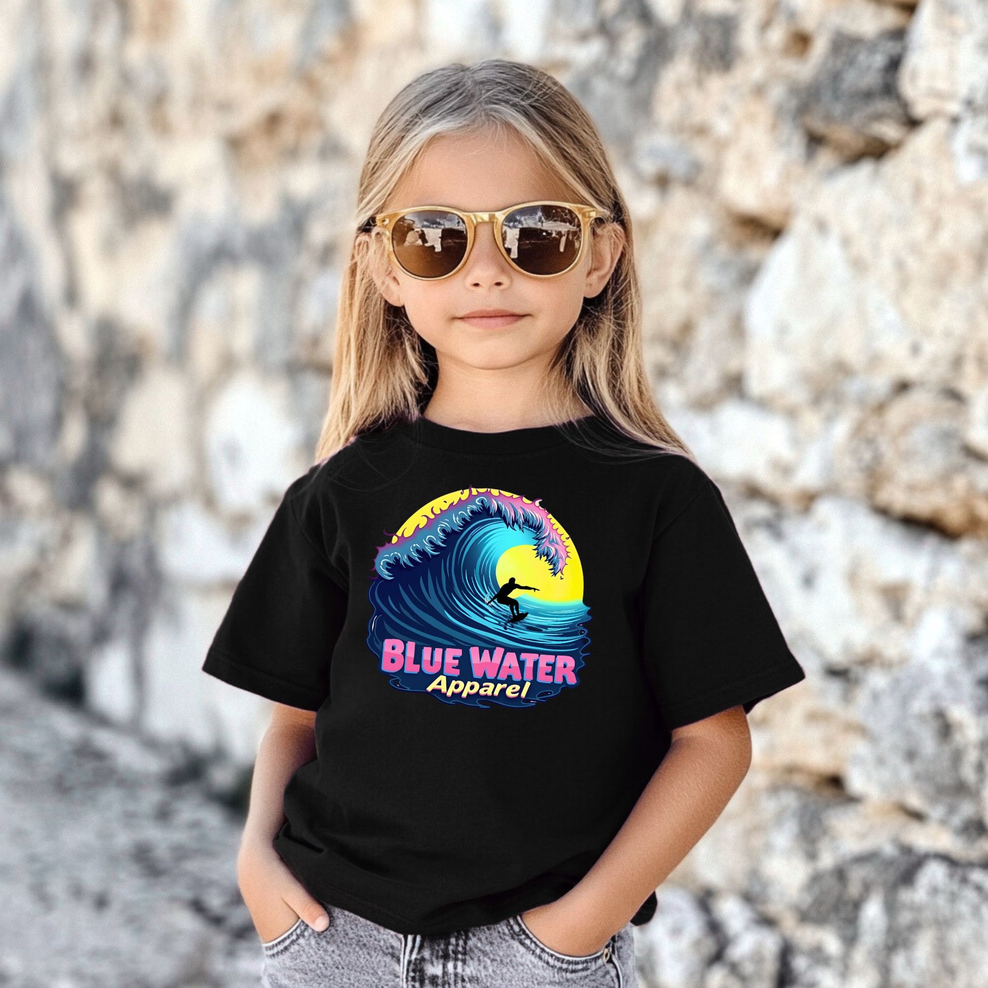 child model wearing black neon surfer tee standing against a stone wall wearing sunglasses by blue water apparel