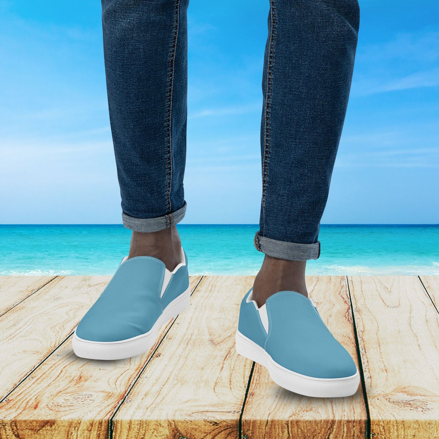 Blue slip-on shoes with white soles worn by a person standing on a wooden platform with a beach background.