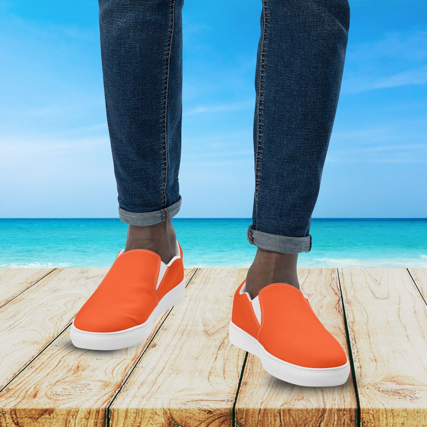 Orange slip-on shoes with white soles worn by a person standing on a wooden platform with a beach background.