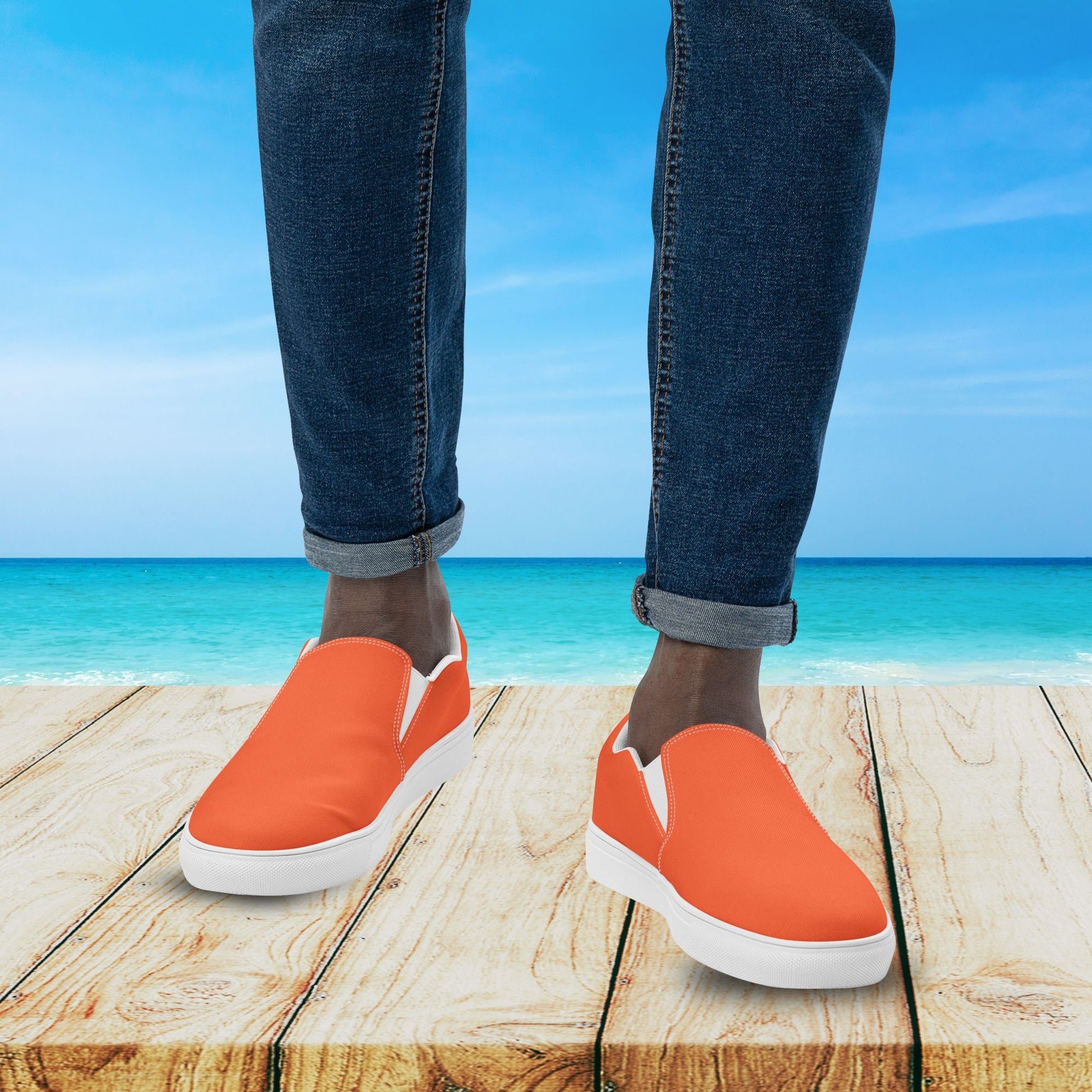 Orange slip-on shoes with white soles worn by a person standing on a wooden platform with a beach background.