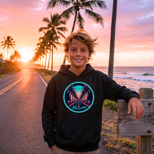 boy wearing a black palm tree road kids hoodie standing on beach palm tree lined road by blue water apparel