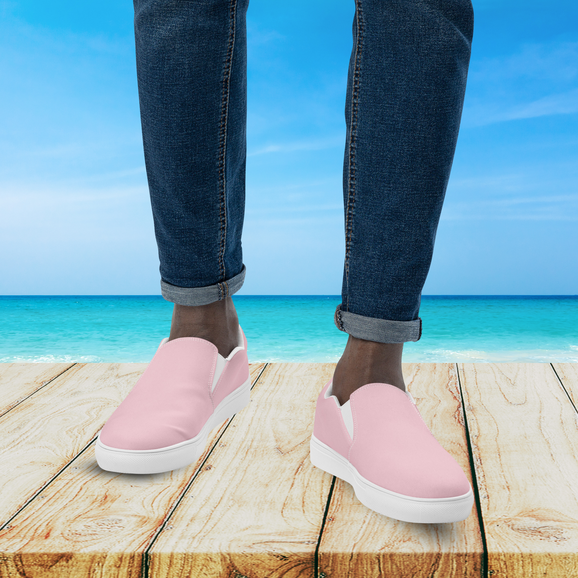 Person wearing pink slip-on shoes with blue jeans standing on a wooden platform by the ocean.