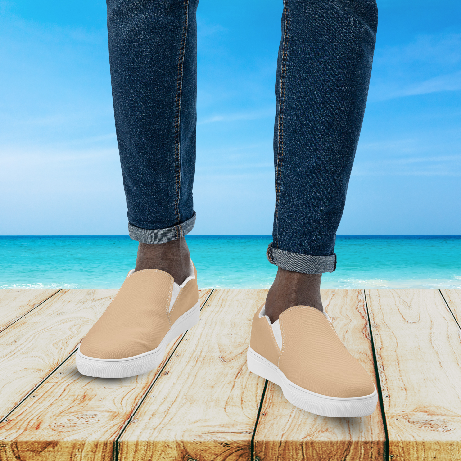 Beige slip-on shoes with white soles worn by a person standing on a wooden platform with a beach background.