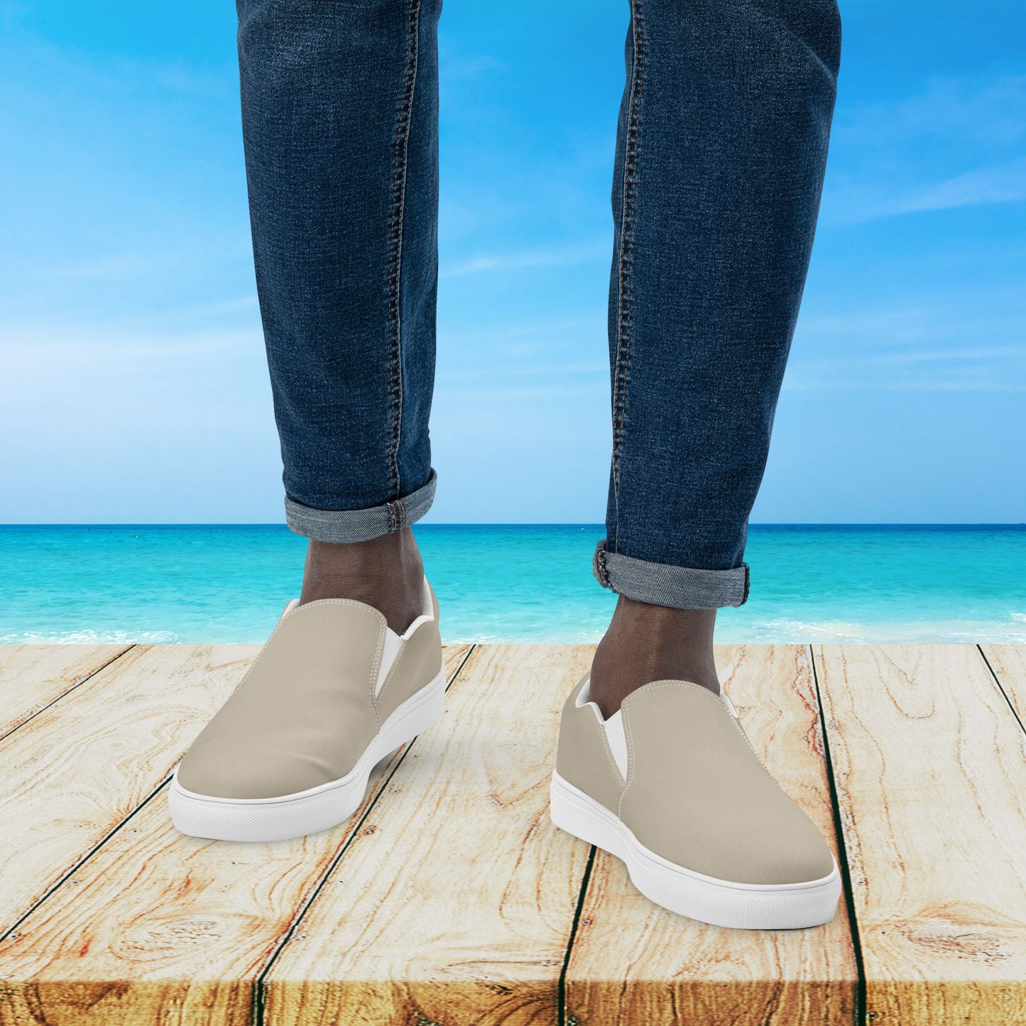 Beige slip-on shoes with white soles worn by a person standing on a wooden platform with a beach background.