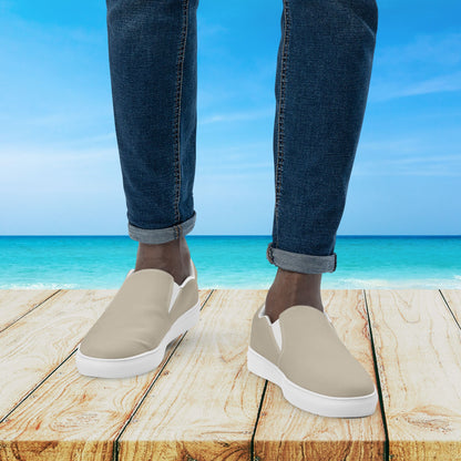 Beige slip-on shoes with white soles worn by a person standing on a wooden platform with a beach background.