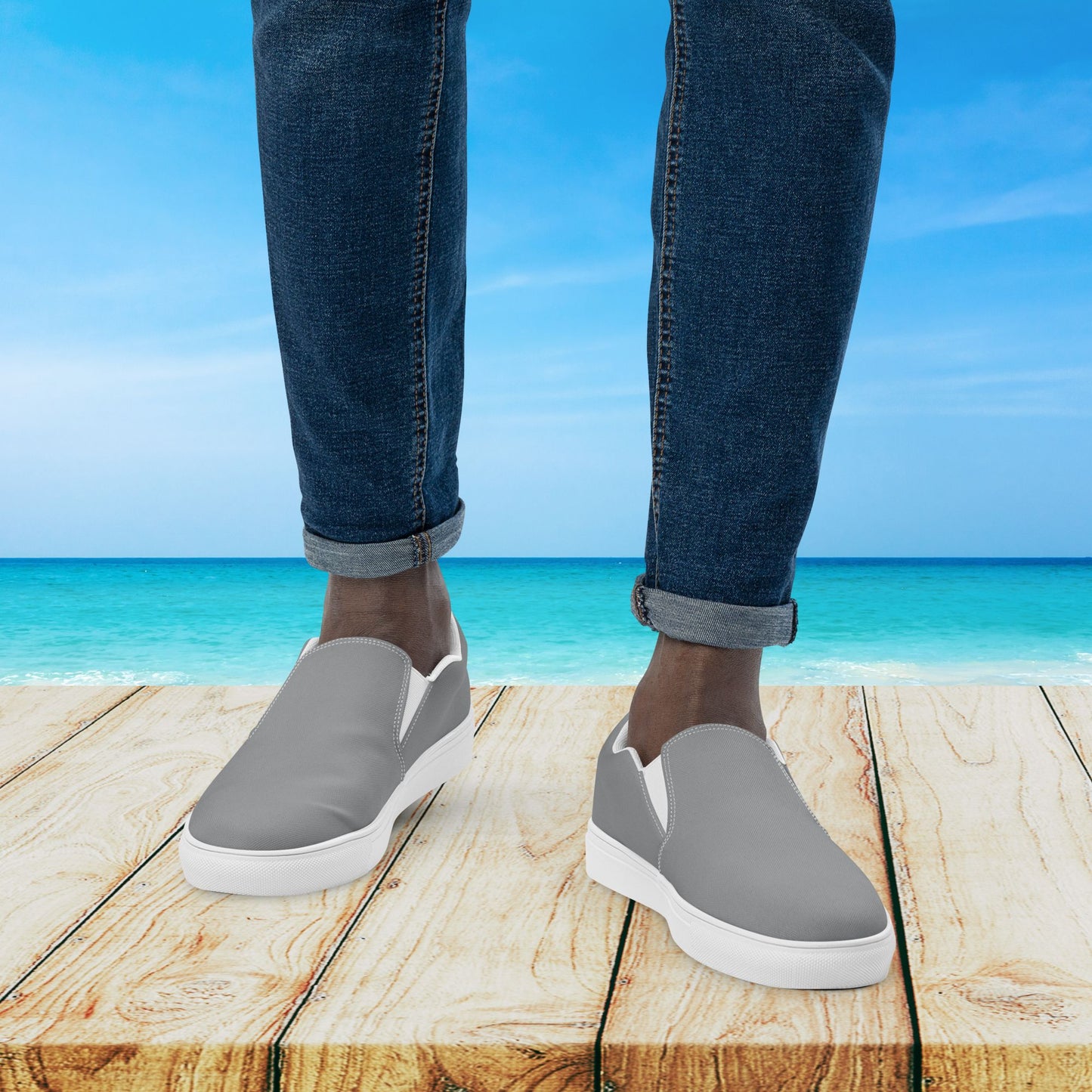 Gray slip-on shoes worn with blue jeans on a wooden platform with a beach background