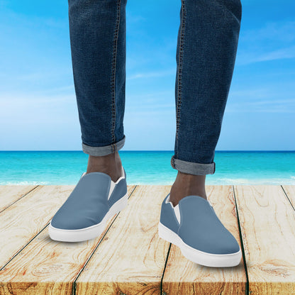 Person wearing blue slip-on shoes with white soles on a wooden platform with a beach background.