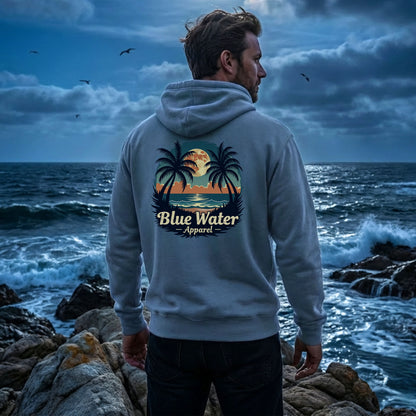 man wearing indigo blue tropical moon adult hoodie standing on rocks of night time beachside by blue water apparel