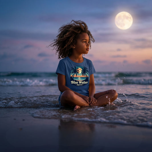 child wearing indigo blue tropical moon kids tee sitting in water on beach by blue water apparel