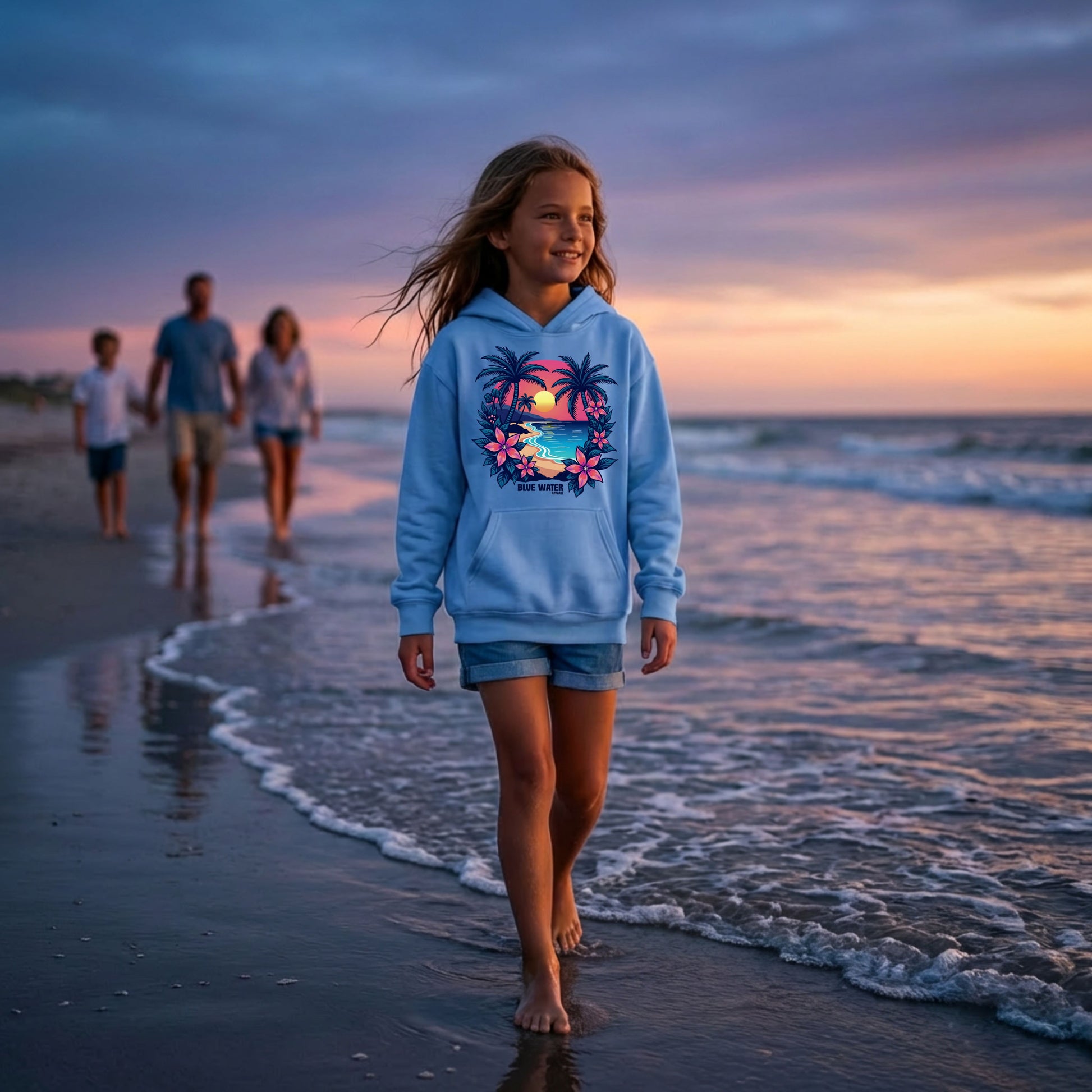 child model wearing carolina blue tropical sunset kids hoodie walking on beach with family in background by blue water apparel