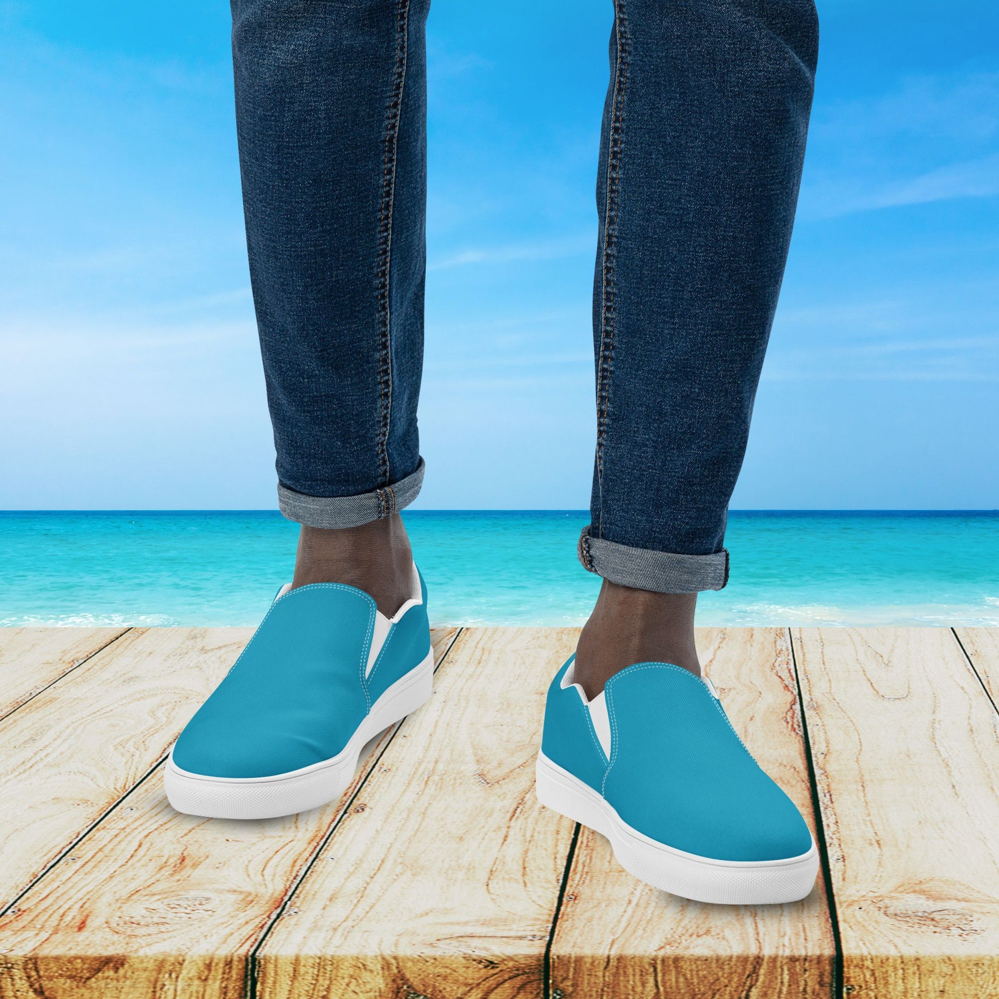 Blue slip-on shoes with white soles worn by a person standing on a wooden platform with a beach background.