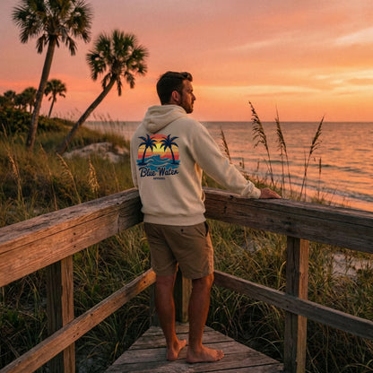 man wearing sand vintage sunset adult hoodie standing on beach pier by blue water apparel