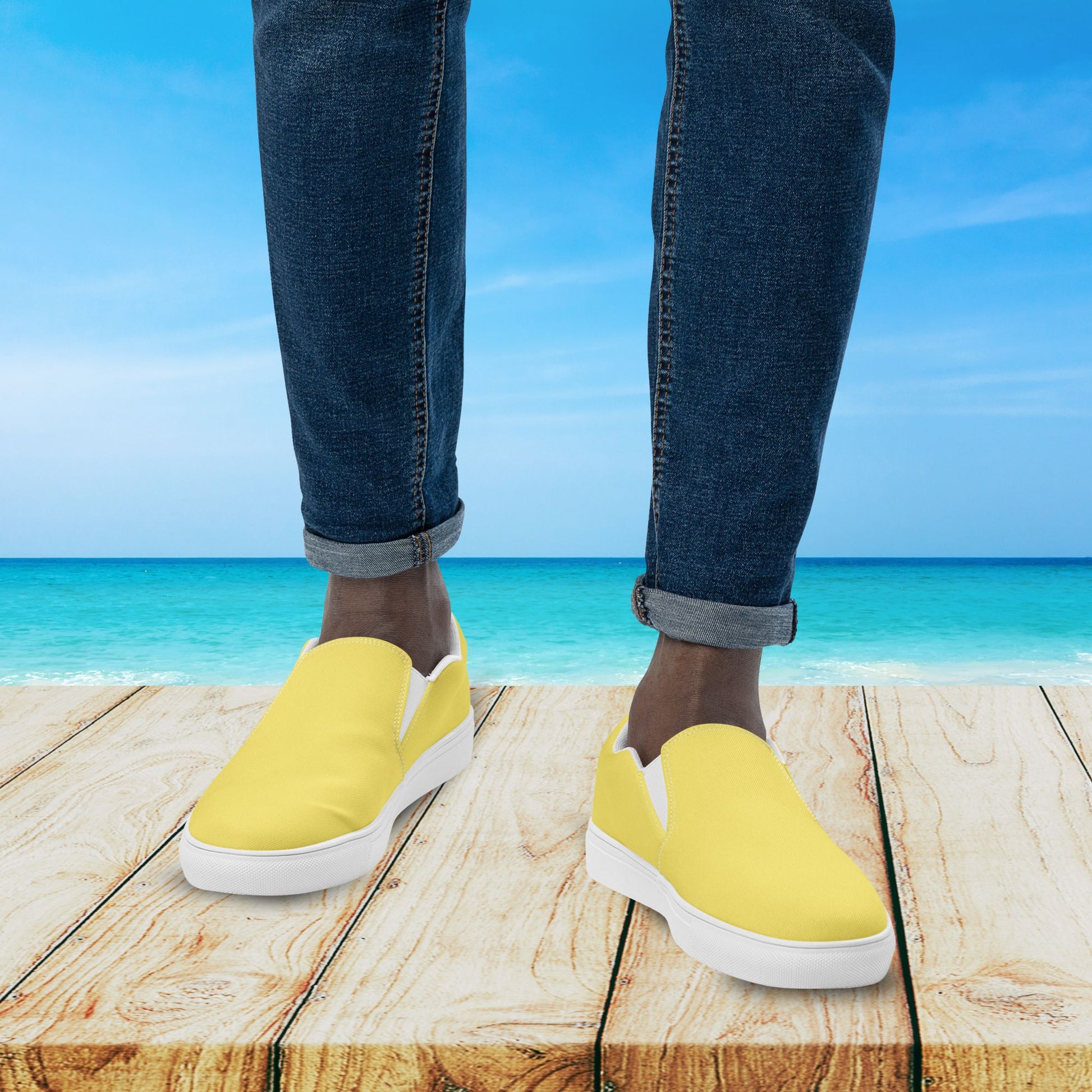 Yellow slip-on shoes worn with blue jeans on a wooden platform with a beach background.